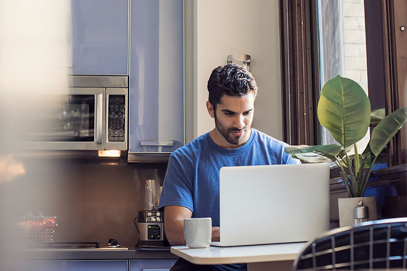 A man sits in his kitchen and works at a laptop computer.