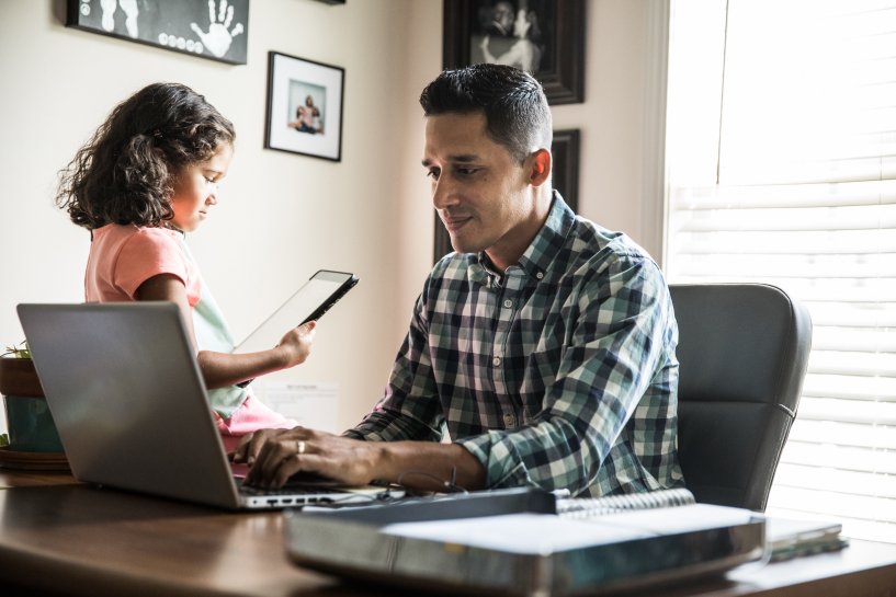 A man works at a laptop while his child sits nearby and plays with a tablet device.